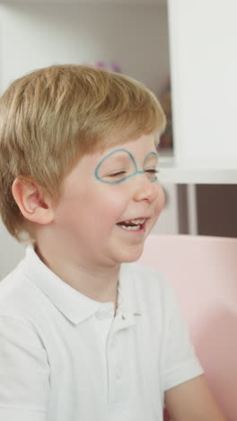 girl paints glasses on younger boy face slow motion. couple of playful children smiles enjoying game. brother and sister play together at home closeup