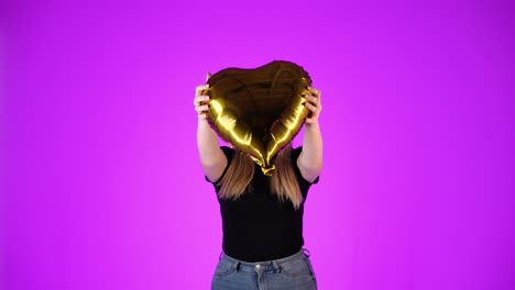 woman in love shows golden heart and smiles, purple background, studio shot