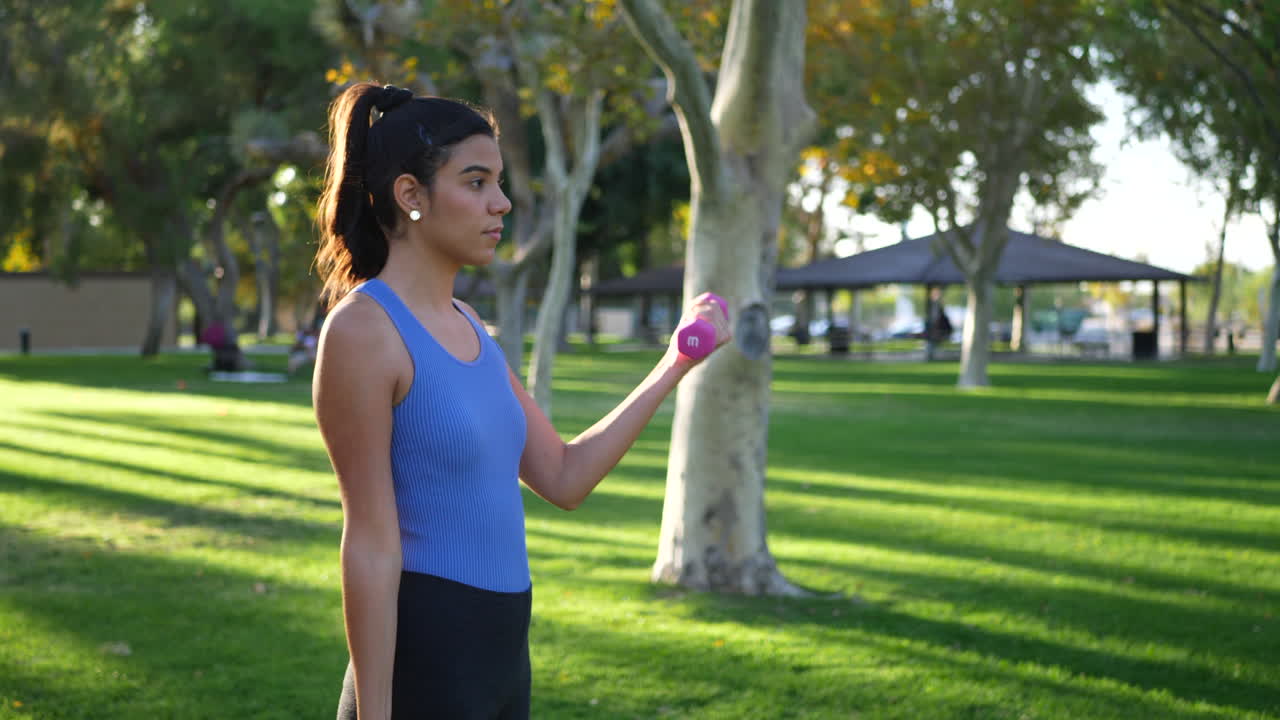 una mujer joven haciendo un entrenamiento con mancuernas en el parque realizando rizos de bíceps para construir músculo del brazo y fuerza a cámara lenta