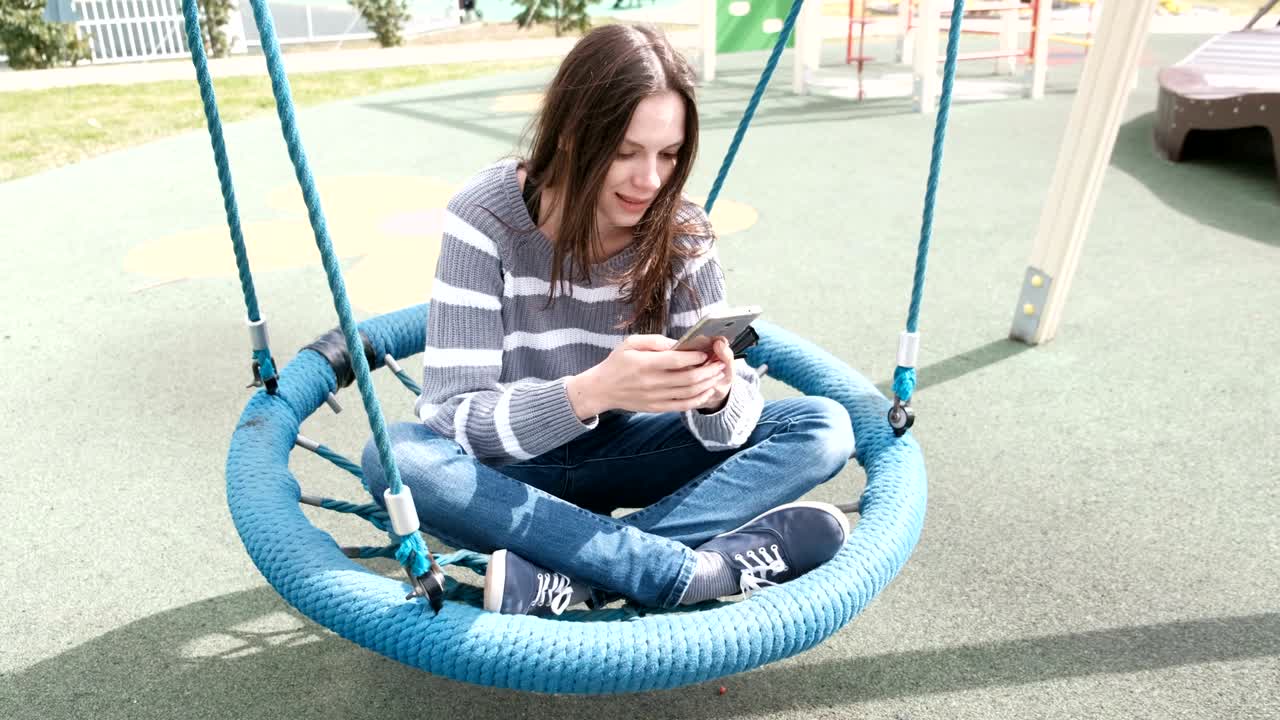 Beautiful brunette woman is swinging on empty webbed swing, loking at social media on mobile phone.