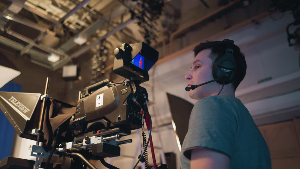 Cameraman in headset operating professional television camera inside broadcast studio while recording presenter on blue screen stage with teleprompter, lighting gear, ceiling rig, and video monitor in frame