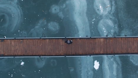 aerial: a man sitting in the middle of a wooden bridge over a frozen lake