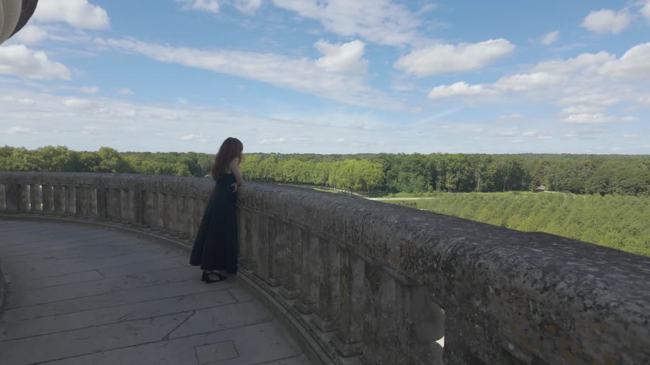 A woman in a long dress stands on the stone balcony of Château de Chambord, overlooking the green forest under a bright summer sky