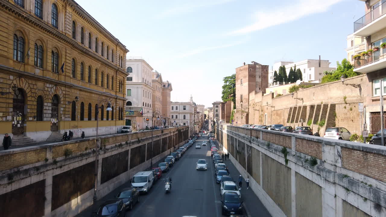 Road the goes towards the colosseum. cars and scooters are passing by and passing under a pedestrian bridge. There are few houses on these of the street and also some roman architecture.