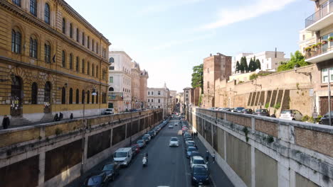 road the goes towards the colosseum. cars and scooters are passing by and passing under a pedestrian bridge. there are few houses on these of the street and also some roman architecture.