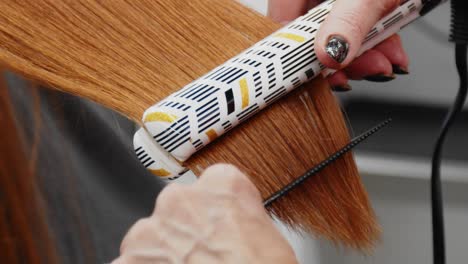 una mujer peinando el cabello rojo de una mujer hacia atrás y luego alisándolo con una plancha alisadora en una peluquería
