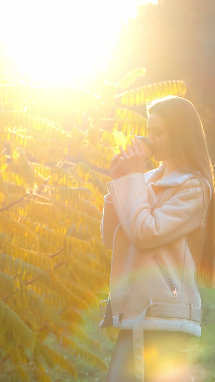 Romantic lady with long dark hair drinking coffee in the park. Woman stopped at the beautiful big bush in the rays of setting sun.