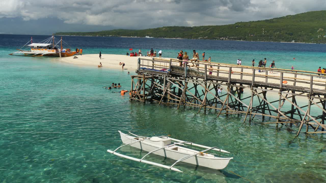 People enjoy a sunny day by the clear waters of Sumilon Island with boats and a wooden pier in view