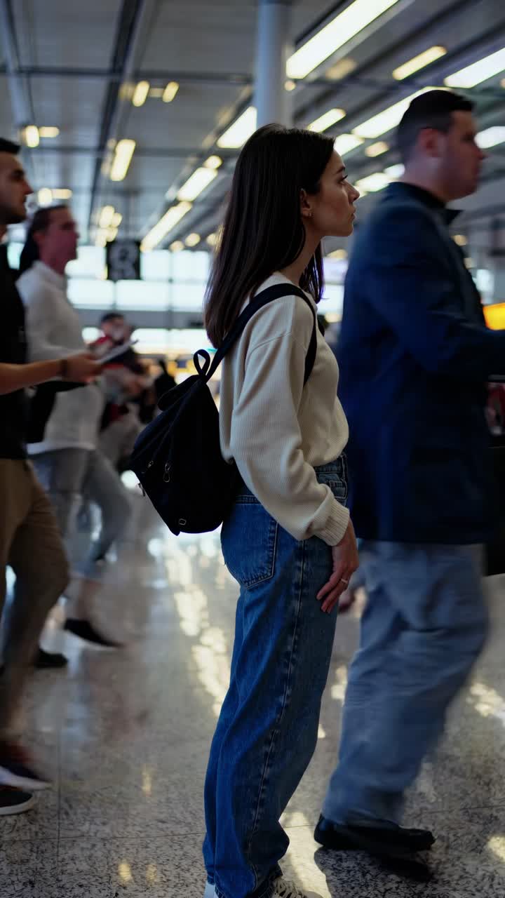 Profil samping seorang wanita di bandara, diambil setinggi mata. Adegan mirip video ini memperlihatkan suasana yang sibuk