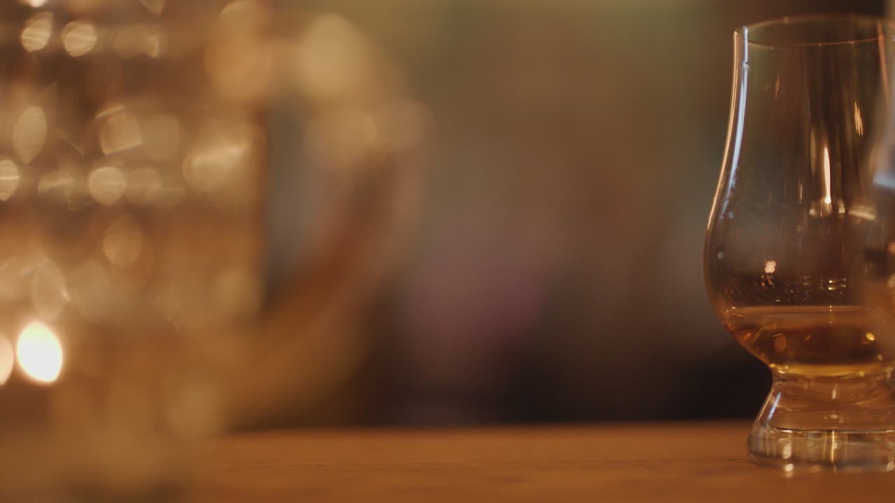 A hand selects a whisky glass from a wooden table in a warmly lit, blurred background bar setting with soft candlelight and shallow focus