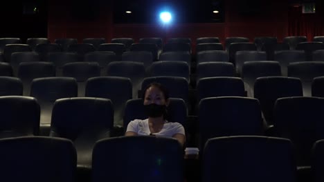 front view of a korean woman sitting alone in the dark cinema and watching movie after a long day with nobody around during covid-19 coronavirus pandemic in south korea wide