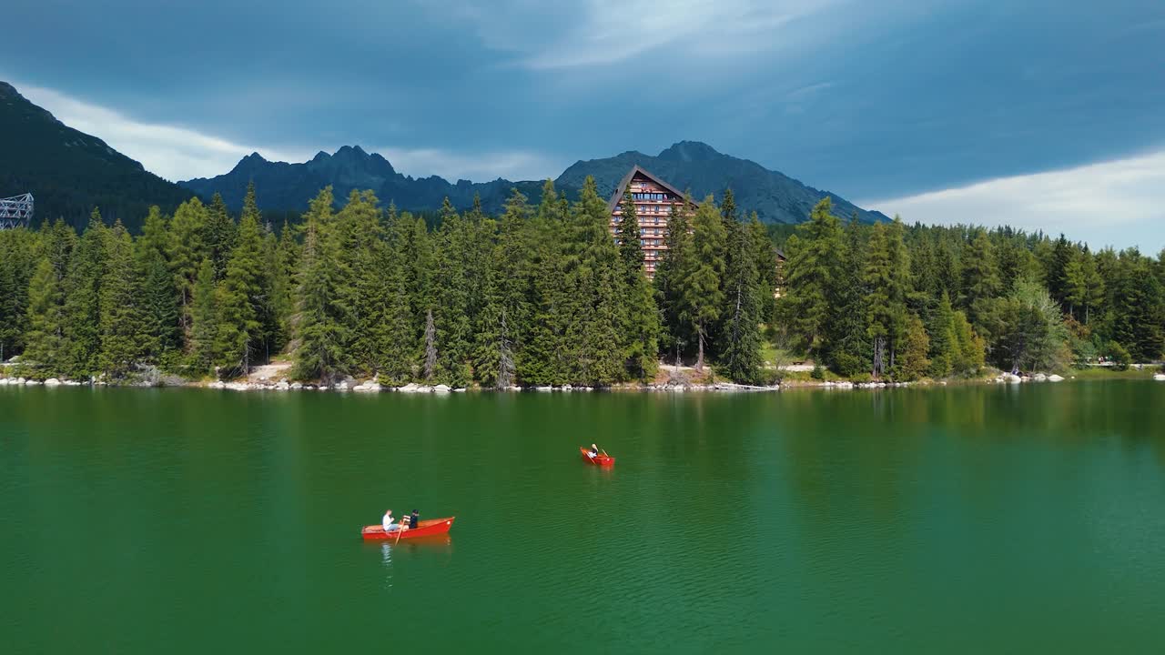 Veduta aerea con drone di barche a remi sul lago Štrbské Pleso in Slovacchia, con le montagne degli Alti Tatra, la riva boscosa e un hotel alpino sotto un cielo drammatico.