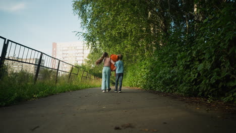 back view of two kids walking together on paved path scattered with dry leaves, bordered by lush green bushes and metal rail fence, distant apartment building visible under blue sky in background