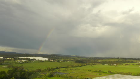 Aerial-view-pans-gently-to-the-left,-showcasing-a-beautiful-natural-landscape-under-a-colorful-rainbow-against-a-backdrop-of-thick-clouds-in-the-sky