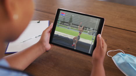 compuesto de una mujer sentada en una mesa con una máscara facial, viendo el evento de salto alto de atletismo en la tableta