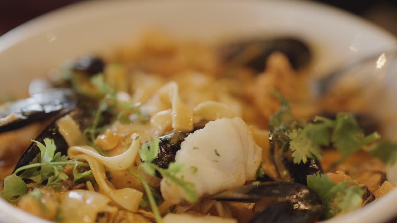 Hand lifts steaming seafood noodle chowder with mussels and herbs in warm, inviting lighting