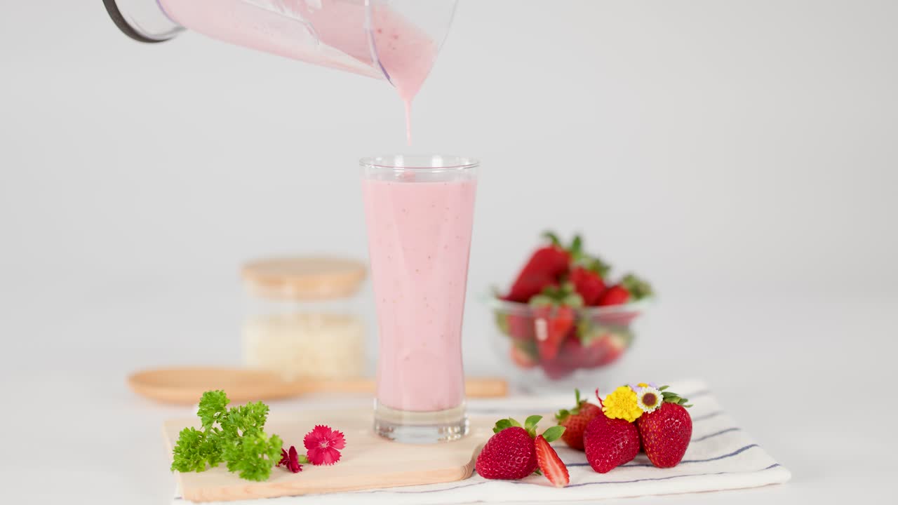 Pink strawberry milkshake pours from blender into glass, surrounded by fresh strawberries, bright lighting