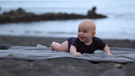 resting on his stomach on the black sand near the ocean, the baby giggles while making eye contact with the camera