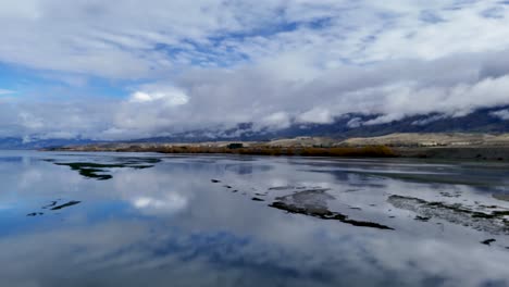drone glides smoothly over tranquil wetlands with water reflections, distant mountains, and dramatic clouds under natural daylight, capturing serene landscape and subtle camera movement