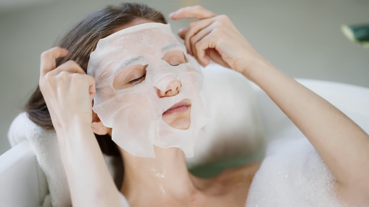 집안의 젊은 여성이 욕조에 누워 있는 동안 피부 관리를 위해 조직 얼굴 마스크를 착용하고 있습니다.