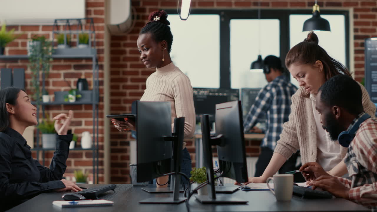 Programer standing up holding digital tablet talking with coder sitting at desk in front of computer screen