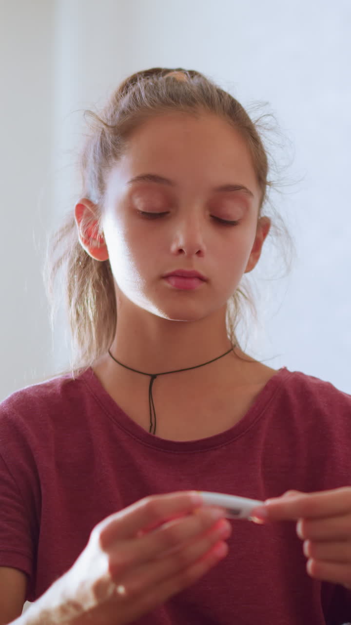 Young girl with ponytail and maroon shirt checks thermometer reading while sitting beside sick person resting off-frame showing care concern and responsibility in calm indoor setting