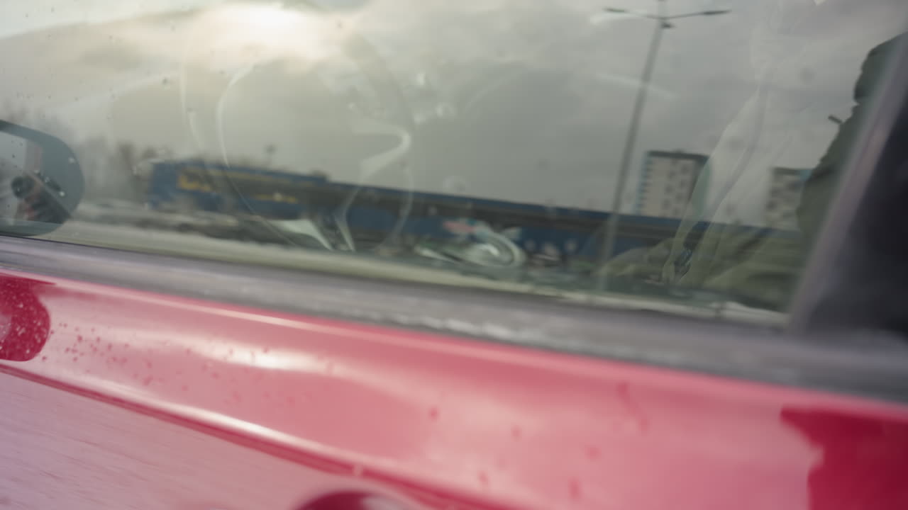 car owner dressed in winter jacket seated inside vehicle during cold season seen rolling up window while facing forward with calm expression, snow-covered trees visible through windshield background