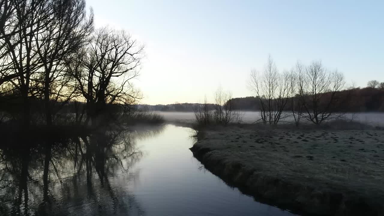 temprano en la mañana, el sol se levanta sobre el pueblo donde un pequeño río fluye a través de la aldea cerca de un campo vacío cubierto de niebla y helada yace en la hierba