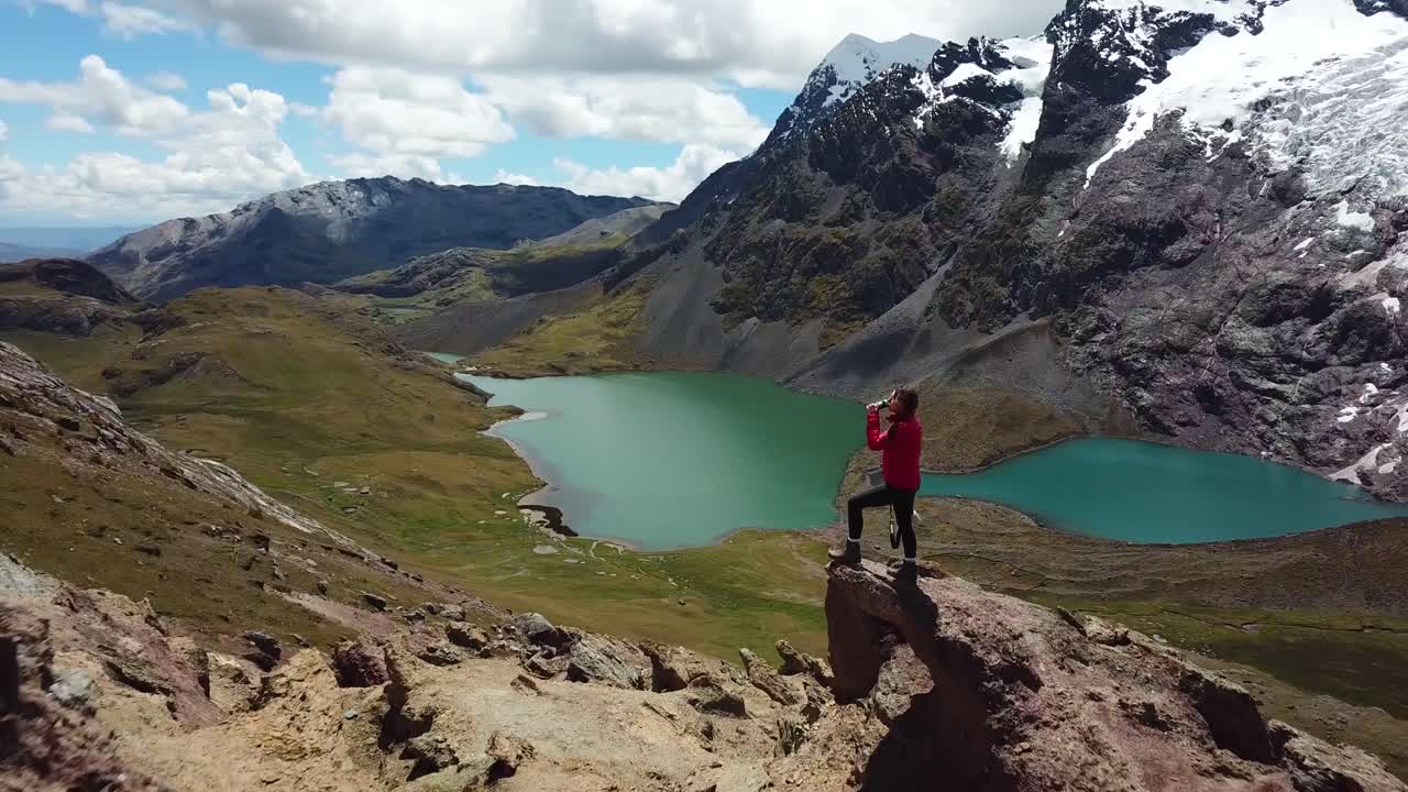 Ripresa aerea con drone sopra una donna che beve acqua, di fronte a laghi glaciali e al ghiacciaio Ausangate, in una giornata di sole, nelle Ande, Perù, Sud America.