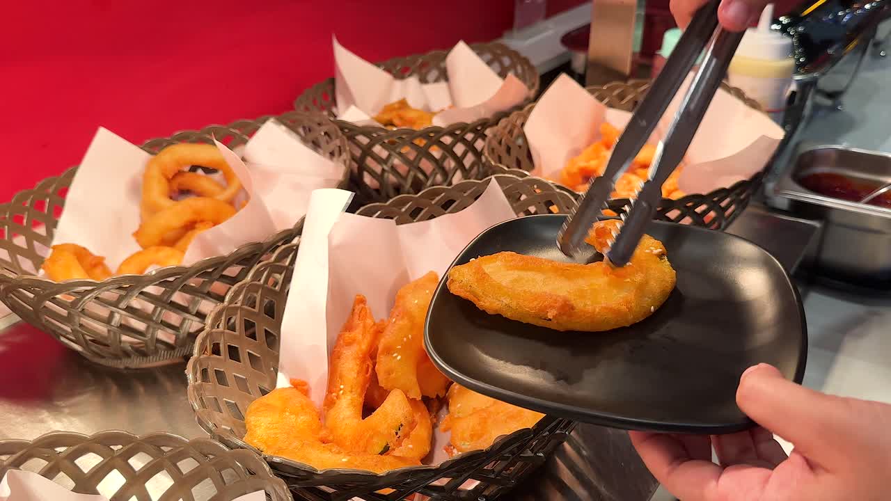 A hand uses tongs to serve golden fried onion rings onto a plate at a Korean BBQ setup