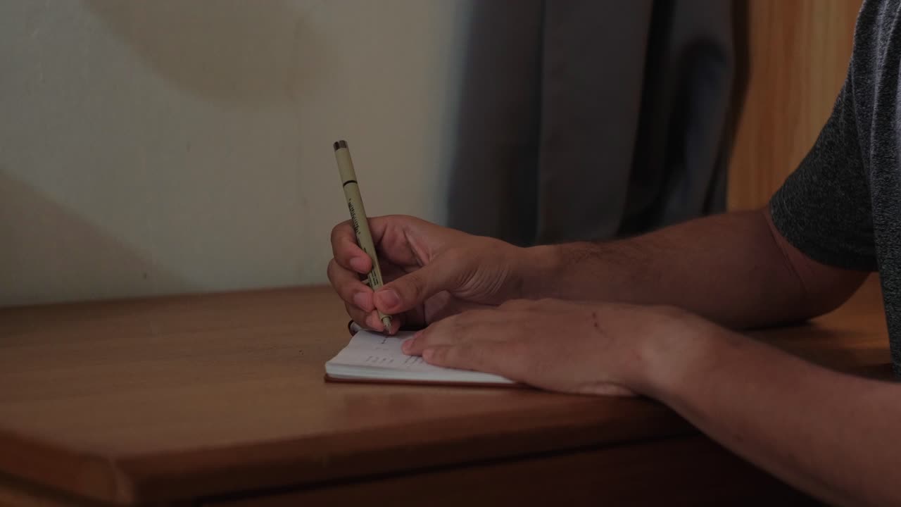 Static medium shot of young man opening a journal notebook and writing inside it, on a wooden desk