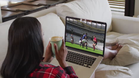 compuesto de una mujer sentada en casa sosteniendo café viendo un partido de fútbol en una computadora portátil