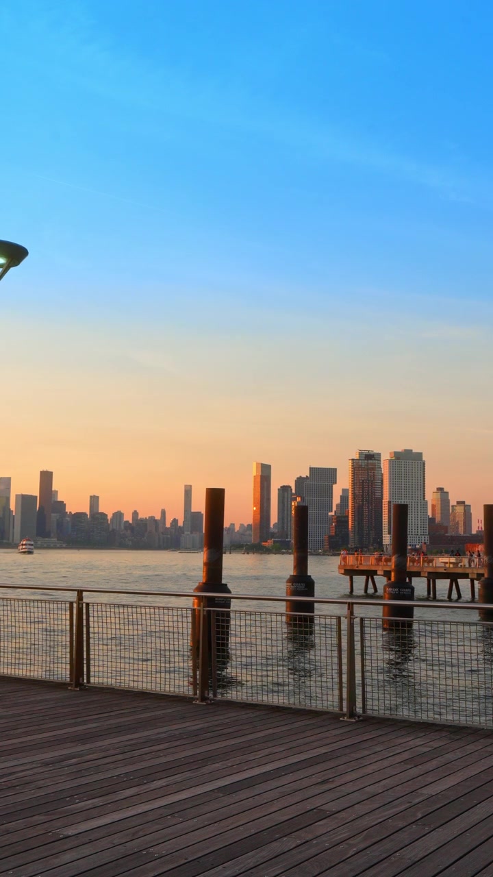 Nova Iorque, EUA, 1 de julho de 2026: Torres à beira-mar de Long Island City ao pôr do sol. Arranha-céus modernos brilham sobre o East River além de estacas escuras e um corrimão de metal.