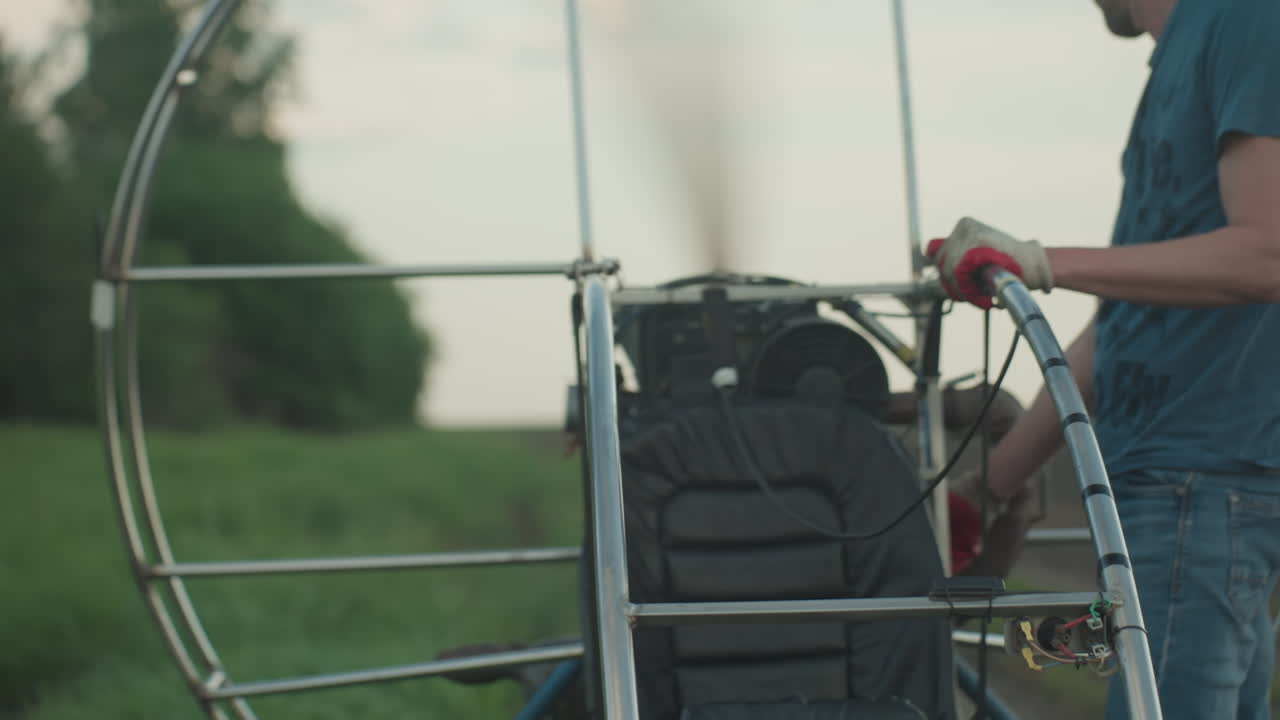 young man in blue shirt with gloved hand pulls throttle on paramotor engine causing fan blades to spin rapidly within circular metal cage on grassy field under pastel sky over open farmland