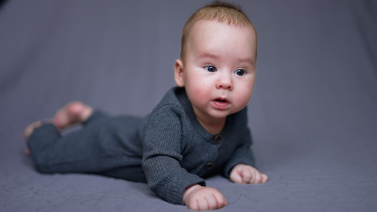 Sweet little boy in grey clothes lies on his belly. Healthy child looks around and then up. Grey blurred backdrop.