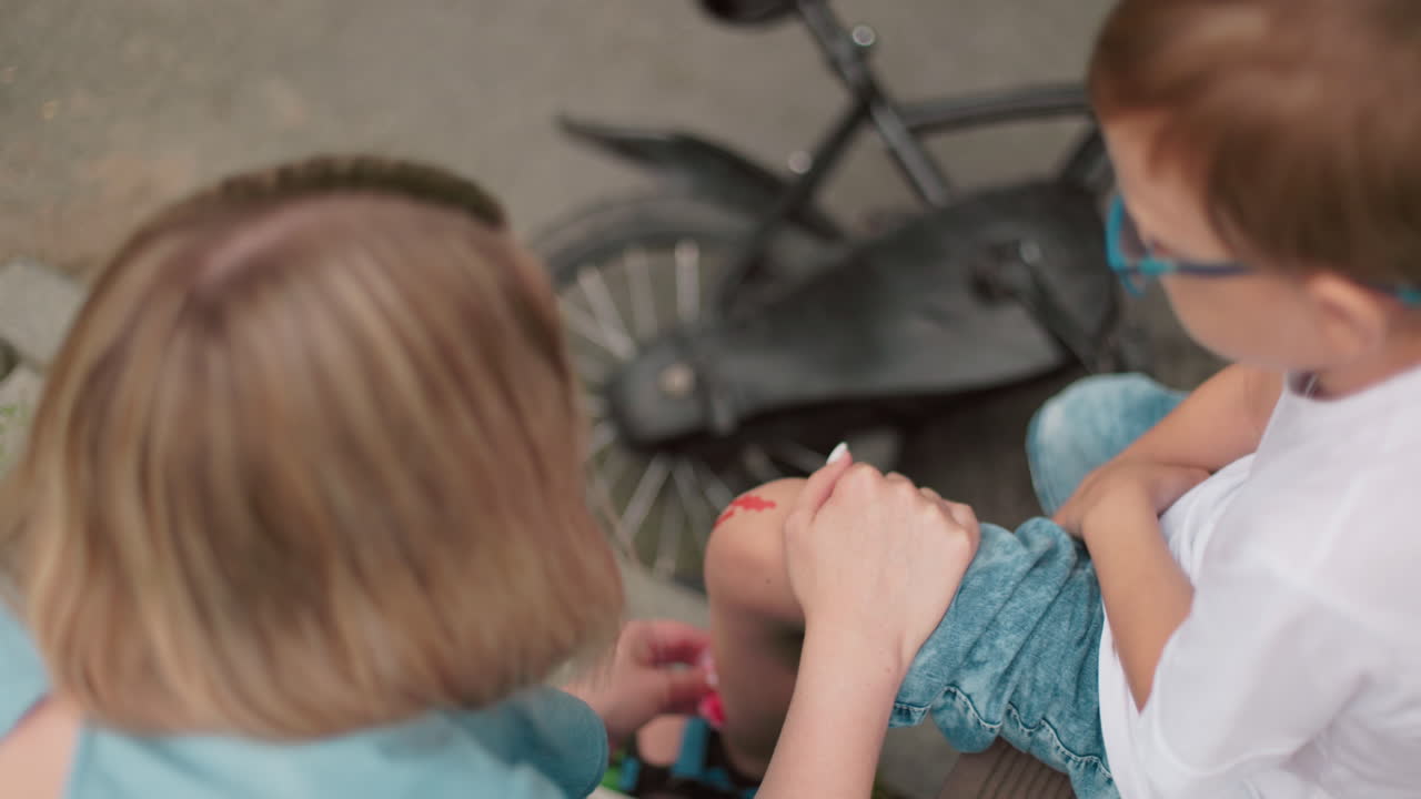 uma mãe ajoelha-se para limpar a perna sangrando de seu filho enquanto sua bicicleta fica no chão ao lado deles