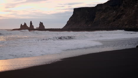 Basaltklippen-Rahmen-Mächtige-Wellen-Ein,-Die-Bei-Sonnenuntergang-über-Einen-Schwarzen-Sandstrand-In-Vik-Island-Krachen