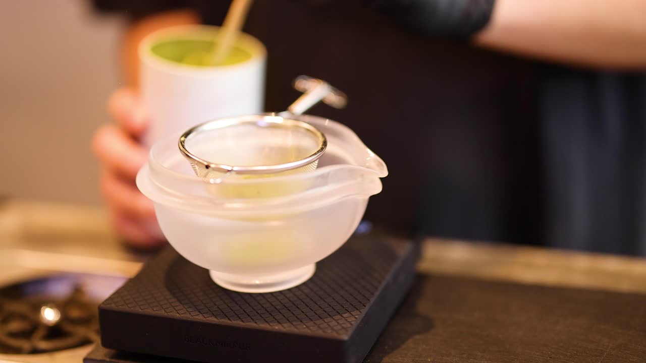 Barista sifts matcha powder into glass bowl under warm lighting, close-up, shallow depth of field