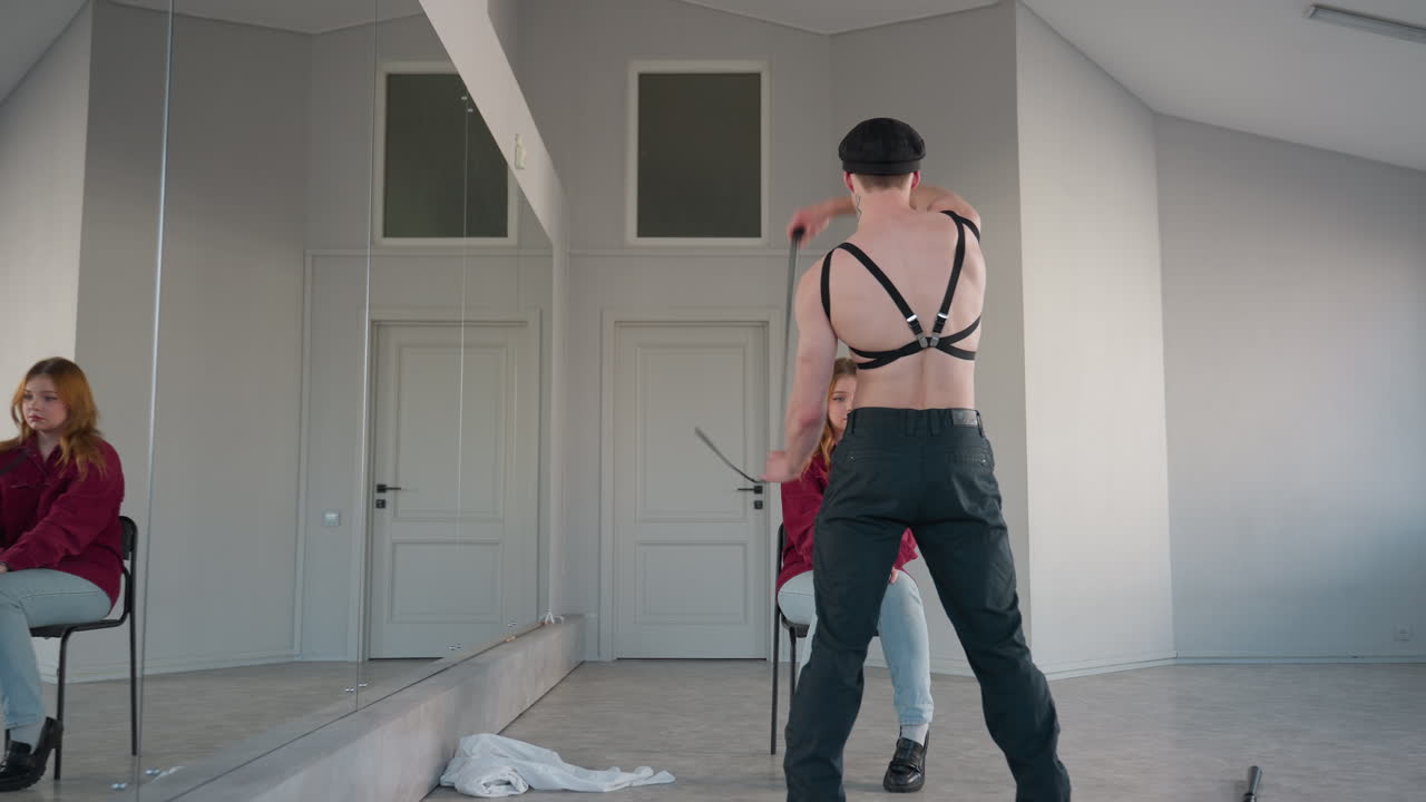 Back view shows shirtless male performer wearing black harness playfully looping belt around neck while dancing before seated woman in red corduroy shirt inside spacious mirror studio
