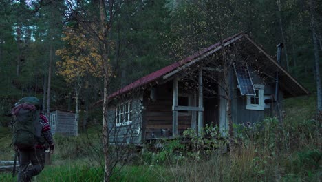 Backpacker-Walking-Towards-Wooden-Cabin-In-The-Forest-In-Trondelag,-Norway