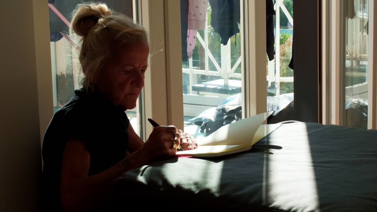 Anziana signora seduta vicino a una finestra, profondamente intenta a scrivere nel suo diario. Luce naturale e soffusa che illumina una scena pacifica di crescita personale, studio o lavoro creativo in un ambiente domestico