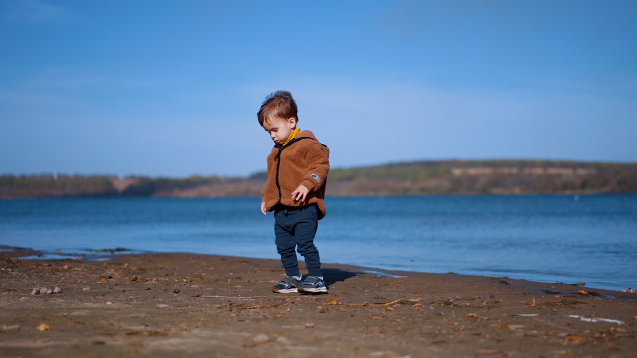 Focused baby boy walks by the river bank. Child finds a stone and takes it to water and throws ahead.