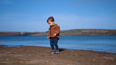 focused baby boy walks by the river bank. child finds a stone and takes it to water and throws ahead.