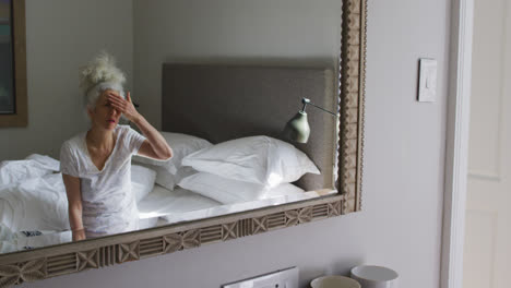 Senior-caucasian-woman-touching-her-face-looking-in-the-mirror-sitting-on-the-bed-at-home