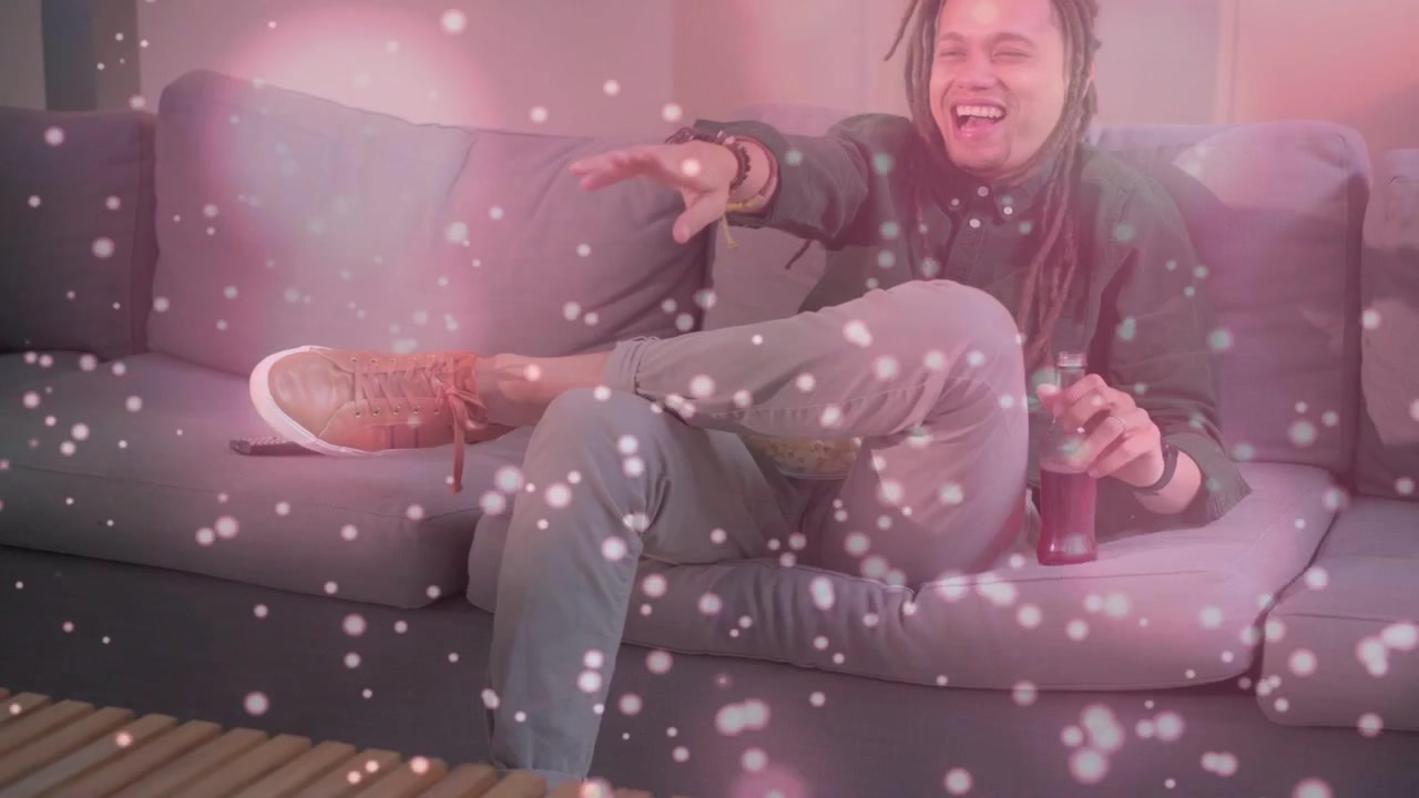 Video verticale: Uomo che preme il telecomando, fa zapping in TV e sorseggia una bottiglia sul divano, con un bokeh rosa che vela la scena.