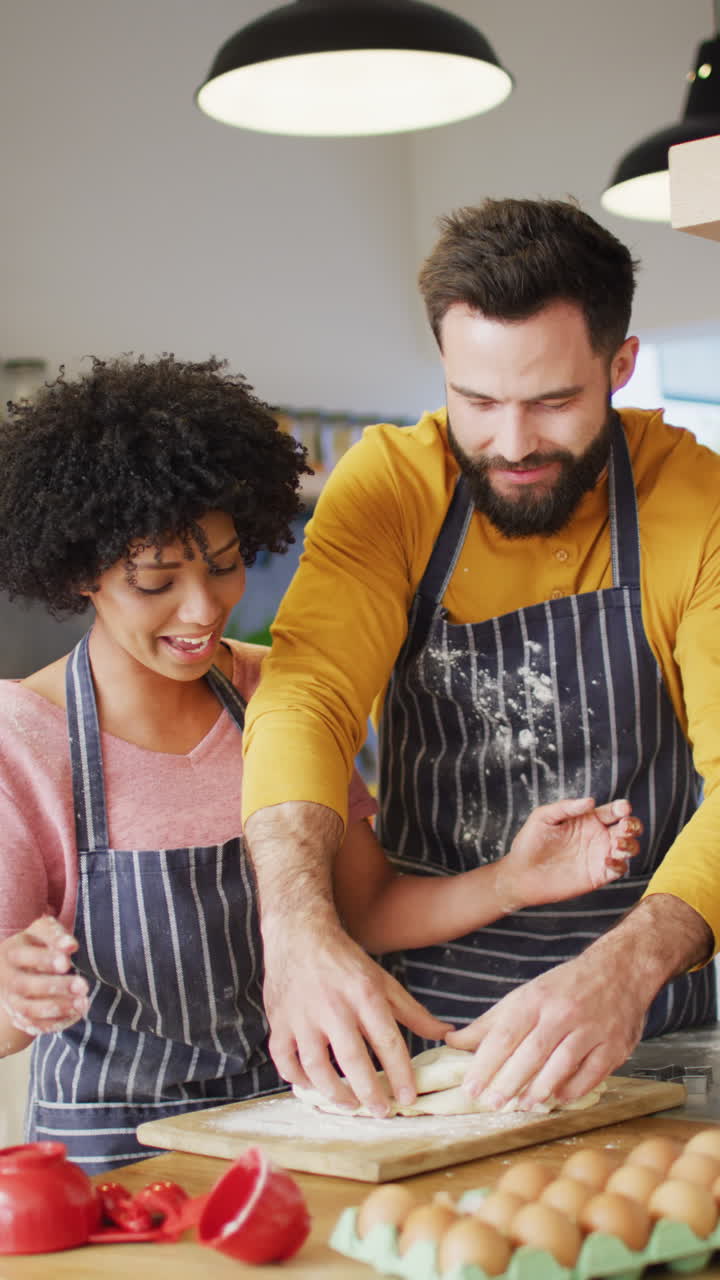Vertical video of happy diverse couple baking in kitchen, kneading dough