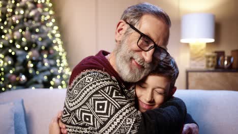 close up of loving happy grey haired grandpa in glasses spending christmas together with joyful grandson sitting in decorated room near glowing xmas tree smiling and hugging on new year eve