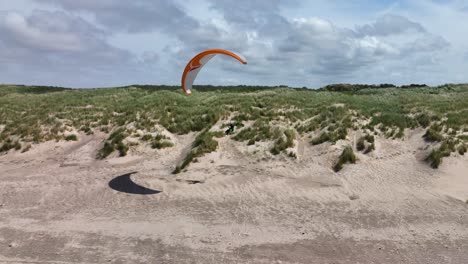 aerial shot of a paraglider riding the lift band of a green grass covered dune with white sand on a sunny day by the sea at ouddorp, brouwersdam, netherlands