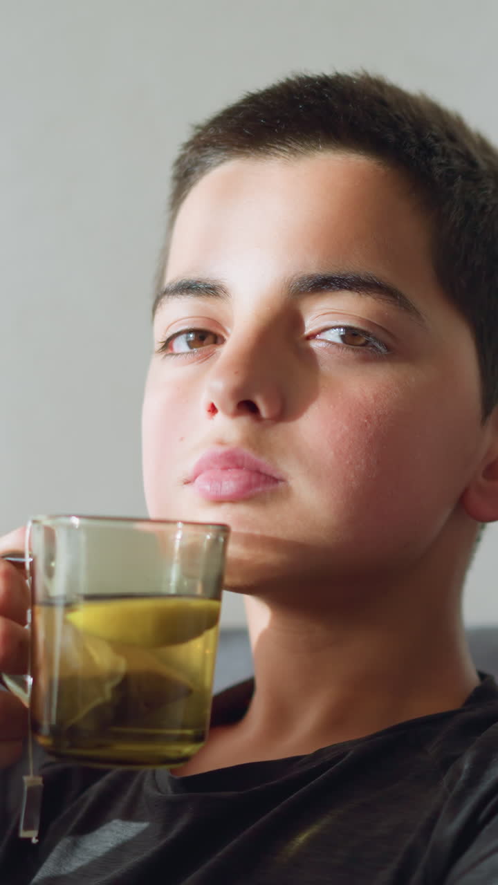 Boy sitting on a couch holding a tea cup close to his mouth, staring directly at the camera with a calm and focused expression, creating a serene and contemplative mood