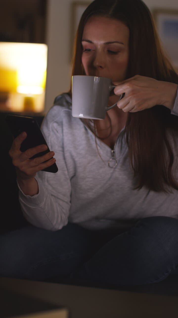 Woman Sitting On Sofa At Home At Drinking Hot Drink And Streaming Or Watching Movie Or Show Or Scrolling Internet On Mobile Phone At Night